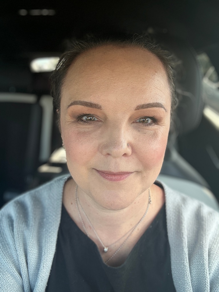 A selfie of a white woman. She is looking straight at the camera smiling. Her eyebrows have been microbladed.
