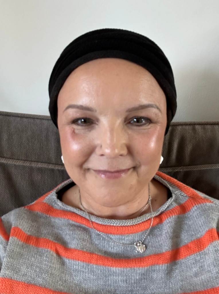 A white woman smiling at the camera. She is wearing a black head wrap to cover her chemo hair loss 